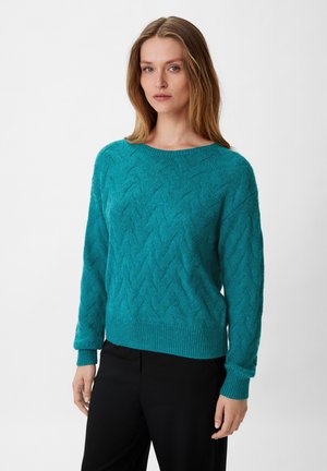 Teal sweater with a zigzag texture, round neckline, ribbed cuffs, and hem. Worn with black pants, features a relaxed fit and soft material.