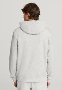 Uomo con capelli castani che indossa una felpa con cappuccio grigia chiara e pantaloni abbinati, girato di spalle su uno sfondo chiaro e semplice.