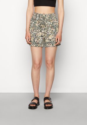 Shorts fleuris avec une taille élastique, fond noir et un mélange de fleurs pastel. Portés avec des sandales noires. Design simple.