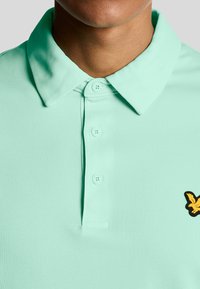 Lichtgroene polo met een klassieke kraag, drie knopen en een geel geborduurd logo op de linkerborst. Gewreven stof.