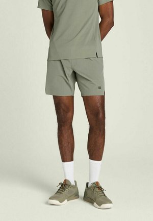 Uomo con pantaloncini sportivi verde oliva, maglietta abbinata, calzini bianchi alla caviglia e scarpe da ginnastica verde oliva, in piedi su sfondo chiaro.