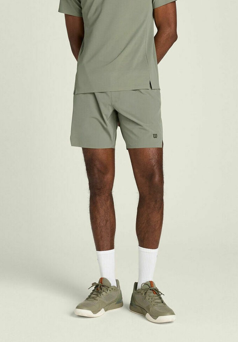 Homme portant un short de sport vert olive, un t-shirt assorti, des chaussettes blanches montantes et des baskets vert olive, debout sur un fond clair.