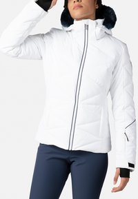 Bílá péřová bunda s vysokým límcem, zipem vpředu a kapucí podšitou námořnickou kožešinou. Obsahuje diagonální prošívání a boční kapsy na zip.