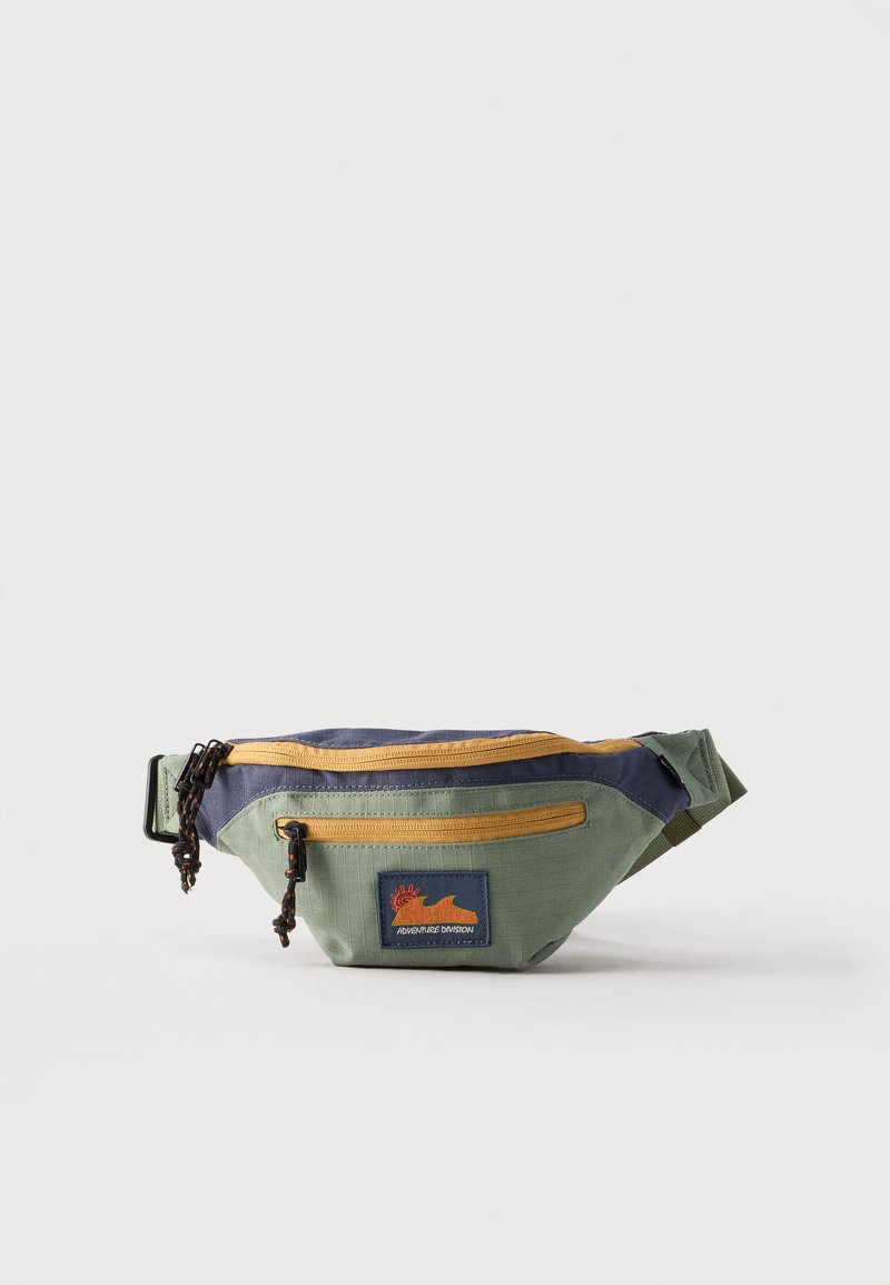 Πράσινο και ναυτικό τσαντάκι μέσης με κίτρινους φερμουάρ, δύο θήκες με φερμουάρ και ένα αυτοκόλλητο με την επιγραφή «Adventure Division».