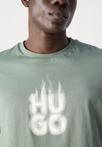 Zelené bavlněné tričko s stylizovaným bílým textem "HUGO" a designem plamene, s kulatým výstřihem a krátkými rukávy.