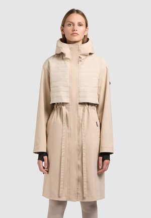 Woman wearing a beige knee-length coat with a high collar, quilted chest panel, drawstring waist, and thumb hole sleeves standing against a plain background.