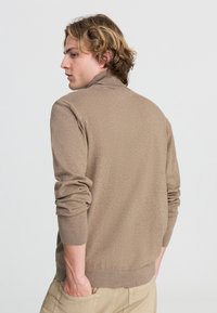 Maglione lavorato a maglia color beige con colletto alto e polsini a coste. Il tessuto sembra morbido, con una texture liscia e una vestibilità casual.