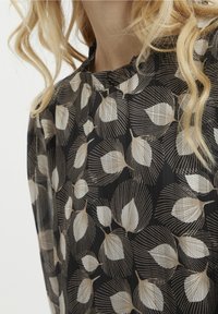 Blouse noire avec un motif de feuilles en blanc et or, en tissu texturé et avec un col froncé. Manches longues et fluides.