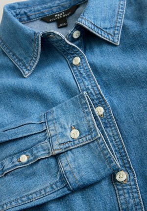 Hellblaues Denimhemd mit klassischem Kragen, Vorderblende mit weißen Knöpfen und Stichdetails. Der Stoff hat eine glatte Textur.