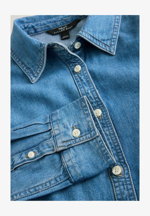 Džínová košile v světle modré barvě, s klasickým límcem, přední légou s bílými knoflíky a švy. Látka má hladkou strukturu.