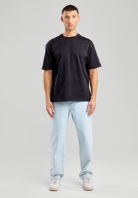 Maglietta nera in cotone con collo a giro, abbinata a jeans leggermente blu e a gamba larga e sneakers bianche, presentata su uno sfondo neutro.