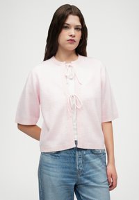 Lichtroze cardigan met korte mouwen, striksluiting en geribbelde details. Zachte textuur, cropped fit, gecombineerd met een witte top en blauwe jeans.