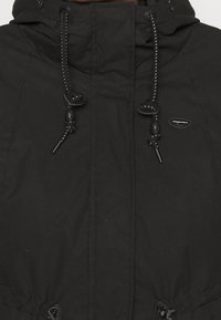 Veste noire avec capuche, cordons de serrage ajustables et détail de logo. Le tissu a une texture mate et une silhouette décontractée.
