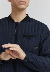 Giacca bomber trapuntata blu navy con colletto a coste nero, zip e tasca sul petto con patch del logo. Dettaglio di cuciture verticali prominenti.