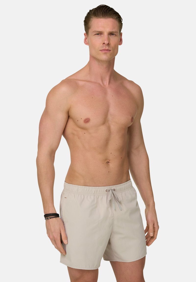 Homme torse nu aux cheveux courts, portant un short de bain beige et des bracelets noirs, debout devant un fond clair uni.