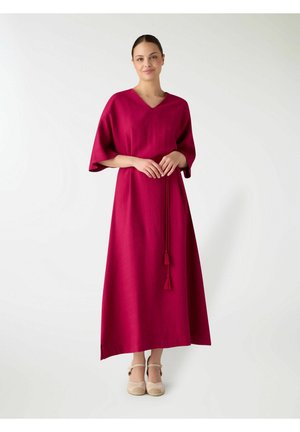 Donna in piedi con un lungo vestito rosso scuro con maniche larghe e cintura con nappine, indossa scarpe beige, su uno sfondo bianco semplice.