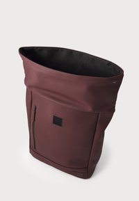 Burgunderfarbene wasserdichte Rucksack mit umklappbarem Deckel, seitlicher Reißverschlusstasche und rechteckigem Etikettenpatch an der Vorderseite. Glatte, matte Textur.