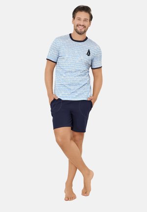 Uśmiechnięty mężczyzna stoi boso z rękami w kieszeniach, ubrany w niebieską koszulkę w paski z motywem żaglówki i granatowe szorty na białym tle.