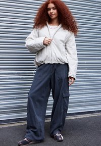 Φουσκωτό μπουφάν ανοιχτό μπεζ με ελαστικό τελείωμα, συνδυασμένο με φαρδιά σκούρα παντελόνια cargo με τσέπες και καφέ παπούτσια με διακριτικές λεπτομέρειες.