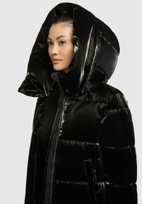 Giacca a vento nera con finitura lucida, cappuccio oversize, design imbottito, zip frontale e tasche laterali, indossata sopra una maglietta nera.