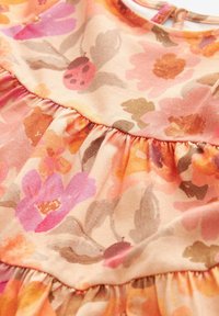 Robe florale en coton doux aux couleurs rose, orange et marron. Elle présente des volants froncés et un col rond avec une fermeture par bouton à l'arrière.
