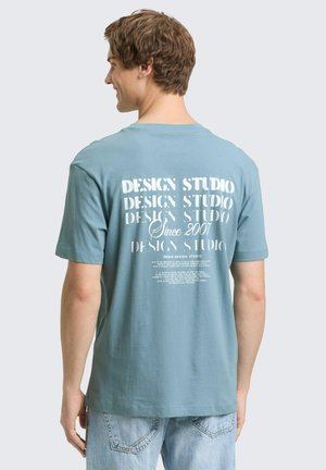 Młody mężczyzna zwrócony tyłem, ubrany w miętową koszulkę z białym napisem "Design Studio Since 2007" oraz jasnoniebieskie dżinsy.