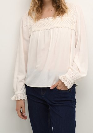 Kobieta ubrana w białą bluzkę z długim rękawem zdobioną koronką na rękawach i dekolcie, w połączeniu z ciemnoniebieskimi dżinsami, stojąca na jednolitym tle.