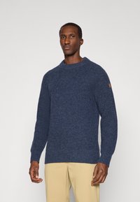 Granatowy sweter z dzianiny o fakturowanym wzorze, z prążkowanymi mankietami i dołem, z małą naszywką logo na lewym rękawie. Noszony z jasnobeżowymi spodniami.