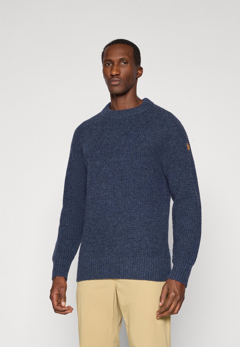 Marine gebreide trui met een textuurpatroon, geribbelde manchetten en zoom, met een klein logo op de linker mouw. Draag met lichtbeige broek.