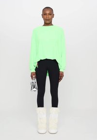 Maglietta lunga oversize verde neon, leggings neri con logo laterale, stivali bianchi alti fino al ginocchio in pelliccia e borsa argentata.