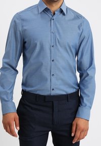 Lichtblauw overhemd met lange mouwen en een button-down kraag, voorzien van een subtiele gestructureerde stof en donkere knopen, gecombineerd met een donkere broek.