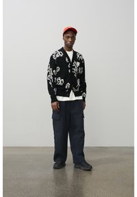 Mladić u crnom kardiganu s cvjetnim uzorkom, bijeloj košulji, mornarskoplavim cargo hlačama, crnim cipelama i crvenoj kapici stoji na poliranom podu uz sivu zidnu površinu.