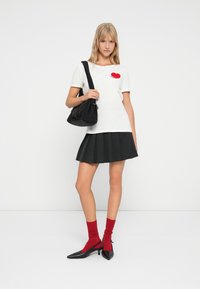 Νεαρή γυναίκα με λευκό μπλουζάκι με κόκκινη καρδιά, μαύρη πλισέ φούστα, κόκκινες κάλτσες, μαύρες γόβες και κρατώντας μια μαύρη τσάντα ώμου, όρθια.