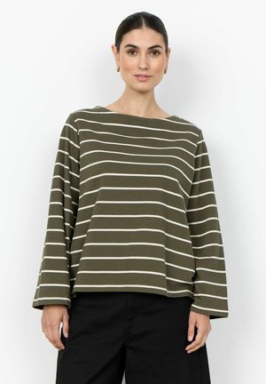 Femme aux cheveux foncés attachés en un chignon bas, portant un haut à manches longues rayé olive et blanc avec un pantalon noir, sur fond blanc.