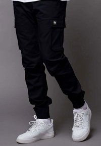 Pantaloni cargo neri con due tasche laterali, polsini elasticizzati e un piccolo patch con logo. Abbinati a scarpe da ginnastica bianche.