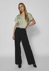 Blusa verde di satin con scollo a V e maniche corte abbinata a pantaloni neri a vita alta e a gamba larga, completati da décolleté nere a punta.