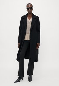 Schwarzer Wollmantel mit strukturiertem Design, kombiniert mit einem beigen Strickpullover, schwarzen Hosen und spitzen schwarzen Stiefeletten.
