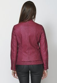 Burgundi faux nahkjakker, millel on tekstureeritud pind, tõmbluku detailid varrukatel, liibuv disain ja kontrastne õmblus seljal.