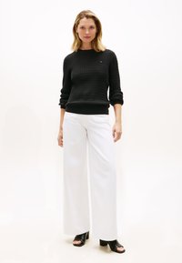 Schwarzer Pullover mit Struktur, weiße Hosen mit weitem Bein und schwarze Sandalen mit Blockabsatz. Das Outfit zeichnet sich durch eine lockere Passform und ein schlichtes Design aus.