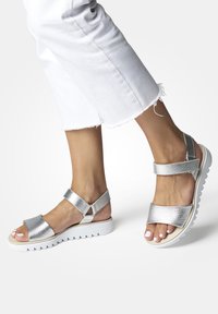 Sandalias metálicas plateadas con un acabado texturizado, dos correas anchas y una suela blanca y estriada. Combinadas con pantalones blancos deshilachados.