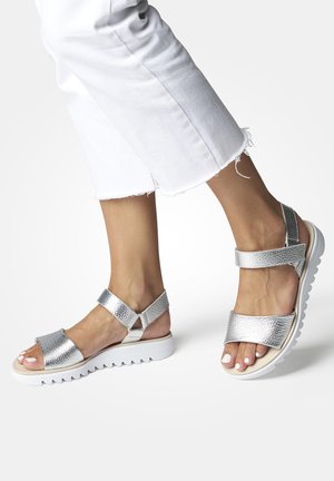 Silver metallic sandals with a textured finish, two wide straps, and a white, ridged sole. Worn with frayed white pants.