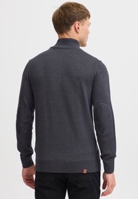 Pull-over gris foncé avec un col montant, des poignets et un ourlet côtelés, et des renforts aux coudes. Fabriqué en tissu tricoté doux, avec un minimum de logo en bas.