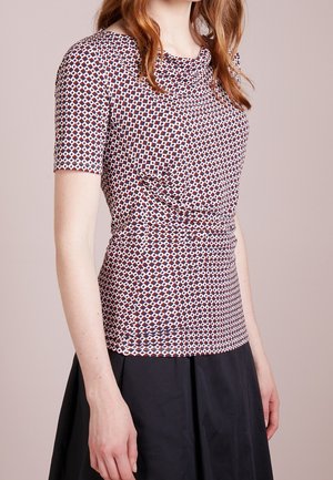 Femme portant un haut à manches courtes à motifs rouges, bleus et blancs avec des formes géométriques, associé à une jupe noire.
