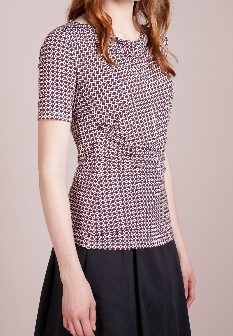 Femme portant un haut à manches courtes à motifs rouges, bleus et blancs avec des formes géométriques, associé à une jupe noire.