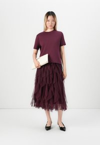 Donna che indossa un top corto a maniche corte di colore viola scuro e una gonna in tulle a strati, con una borsa a pochette bianca, in piedi di fronte a uno sfondo bianco semplice.