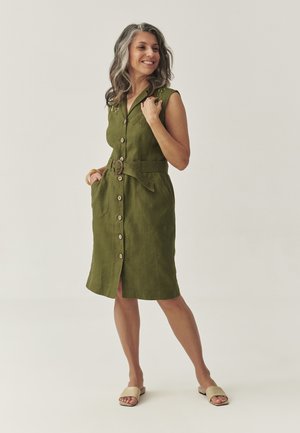 Grünes Leinen-Kleid ohne Ärmel mit Knopfleiste, selbstgebundenem Gürtel und zwei seitlichen Taschen. Flache Sandalen mit offener Zehenpartie runden das Outfit ab.