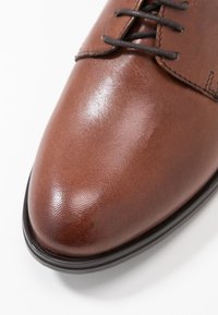Chaussure habillée en cuir marron avec une finition brillante, bout rond, design à lacets et semelle en caoutchouc noir. Accents cousus le long des bords.