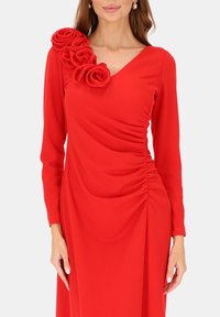 Czerwona sukienka z długim rękawem, z dekoltem w kształcie litery V, marszczeniem po boku i dekoracyjnym bukiecikiem z róż z materiału na ramieniu. Gładka tekstura.