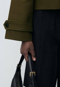 Dark-skinned hand holding black leather handbag with gold buckle, wearing olive green sleeve with black button and black pants.