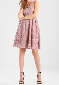 Vaaleanpunainen pitsimekko, jossa on istuva yläosa, aaltoilevat reunat ja A-linjainen helma. Mekossa on ohuet olkaimet ja monimutkaiset kukkakuvioinnit. Mustat korkokengät.